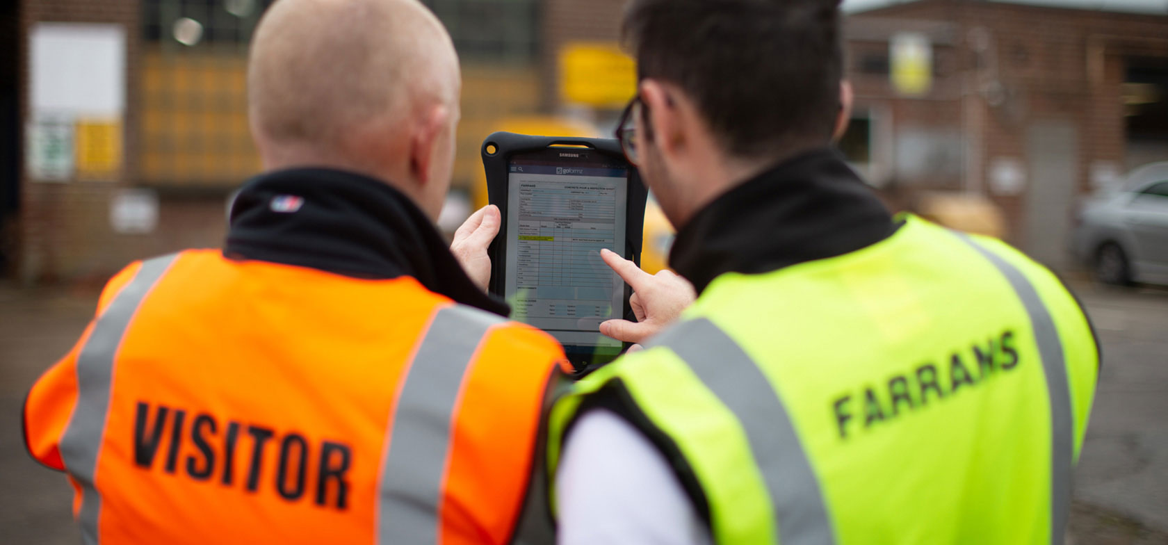Two Farrans Construction engineers fill out a mobile form in the GoFormz app on a tablet, wearing yellow and orange safety vests