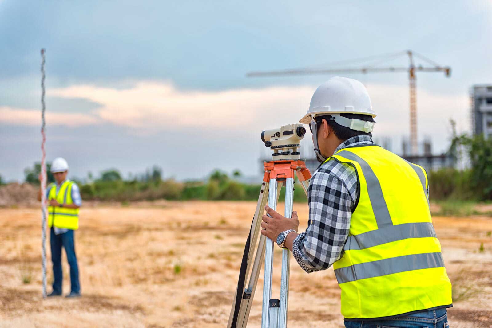 Two land surveyors wear yellow safety vests and hardhats while using land surveying equipment in the field