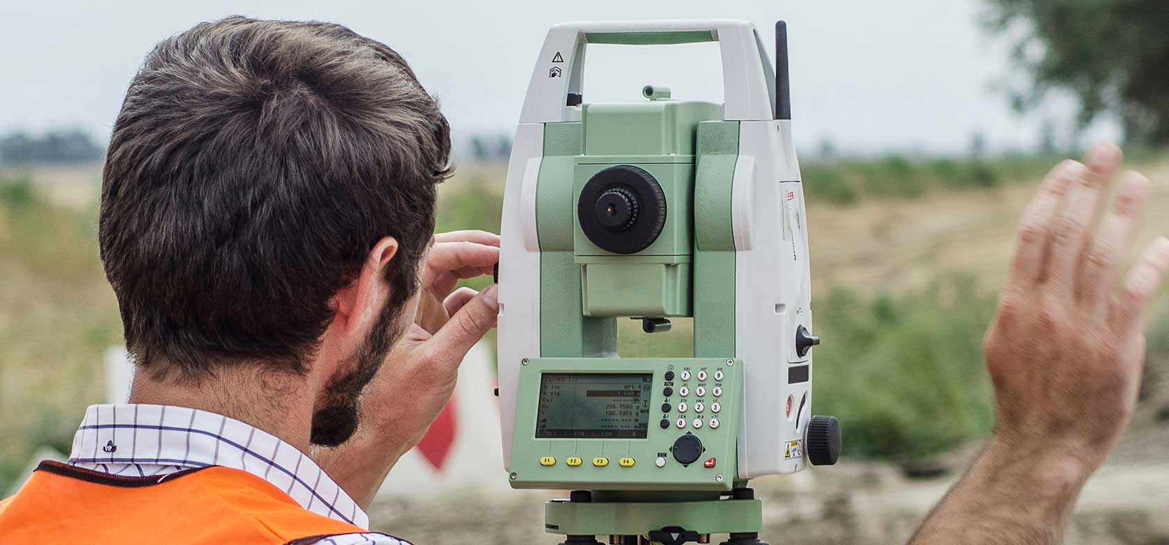 A land surveyor uses surveying equipment in the field wearing an orange safety vest